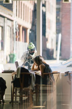 a woman inside a coffee shop looking at her laptop with a man behind the glass looking at his phone in downtown flagstaff