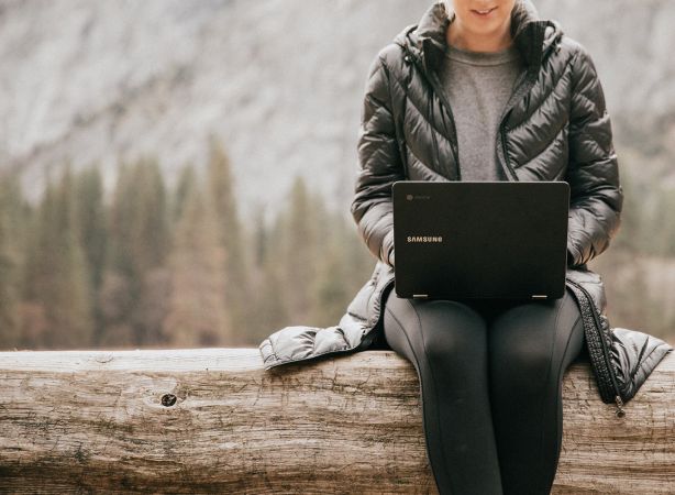 a woman sitting on a log with a laptop in her lap with a mountain behind her